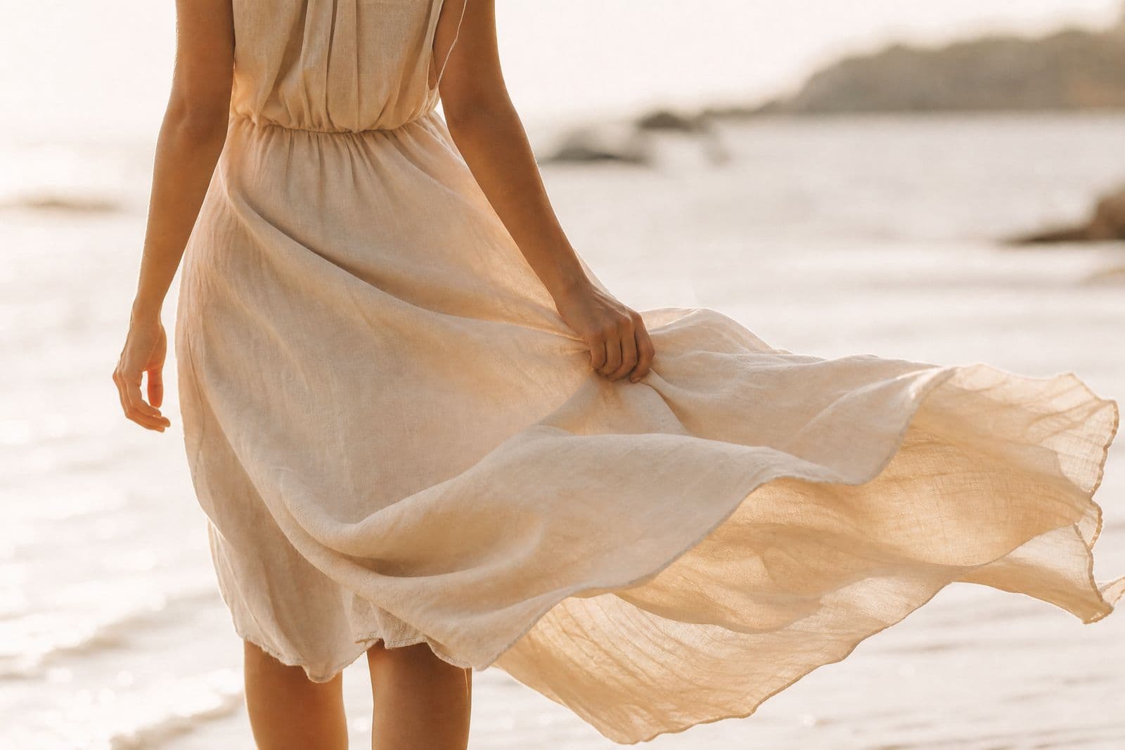 A long linen dress at the water's edge with the hem lifting in the breeze, photographed from the shoulders down.